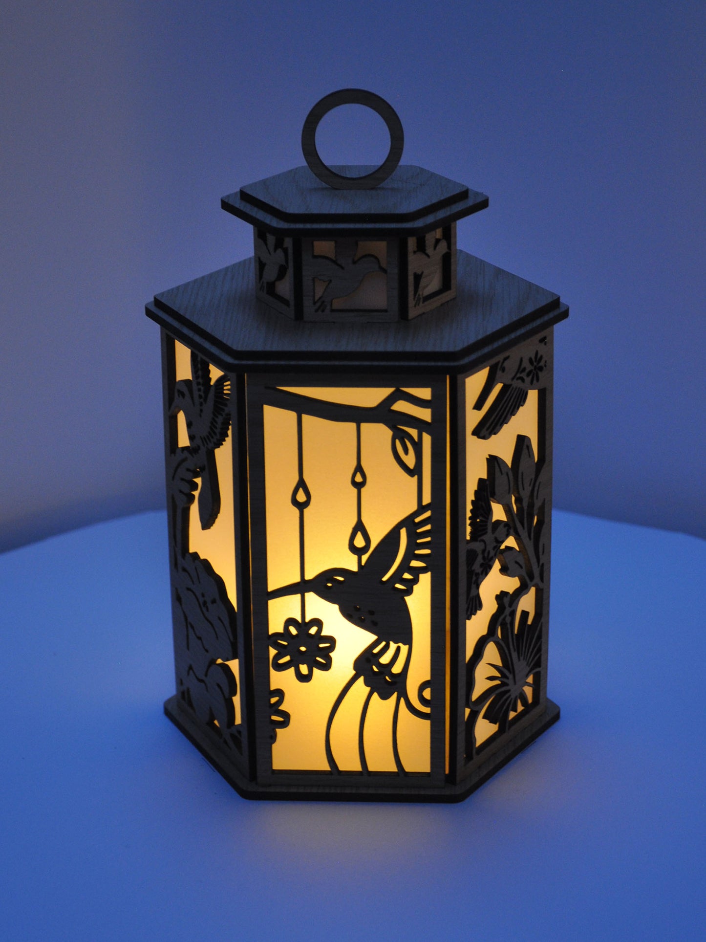 Decorative lantern with cut-out designs of a hummingbird and flowers, illuminated from within.